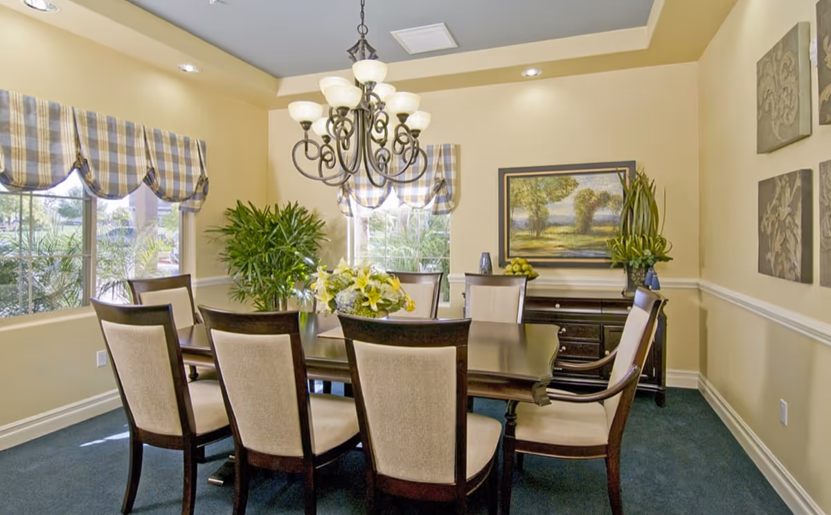 A dining room with a dark wooden table surrounded by eight upholstered chairs. The room has large windows with plaid valances, a chandelier hanging above the table, and decorative plants and artwork on the walls.