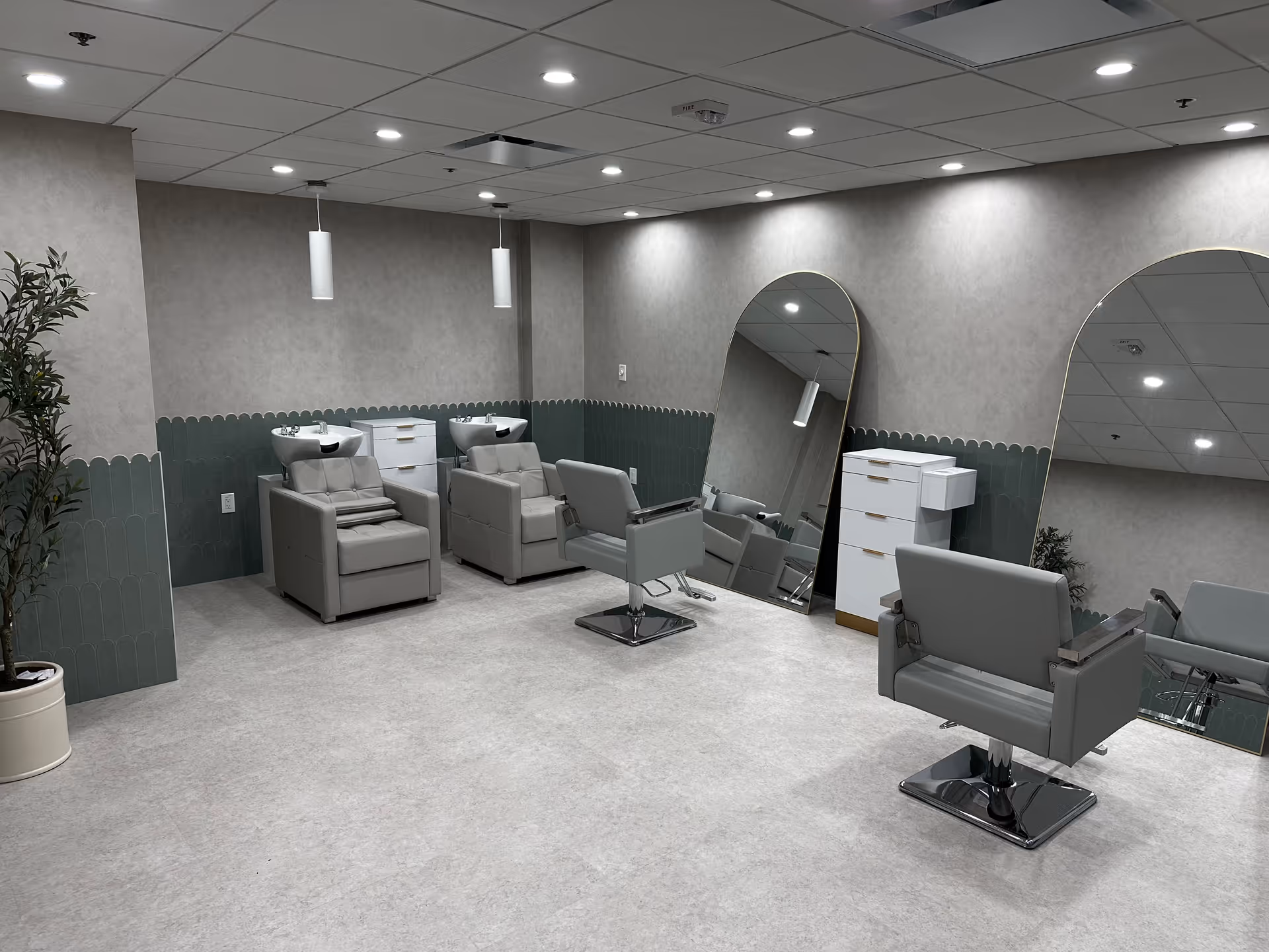 Interior of a modern salon area in a senior living facility with two gray shampoo chairs and sinks, two gray styling chairs, large arched mirrors, a white storage cabinet, and a potted plant in the corner. The walls have a two-tone design with gray and green scalloped paneling, and the ceiling has recessed lighting.