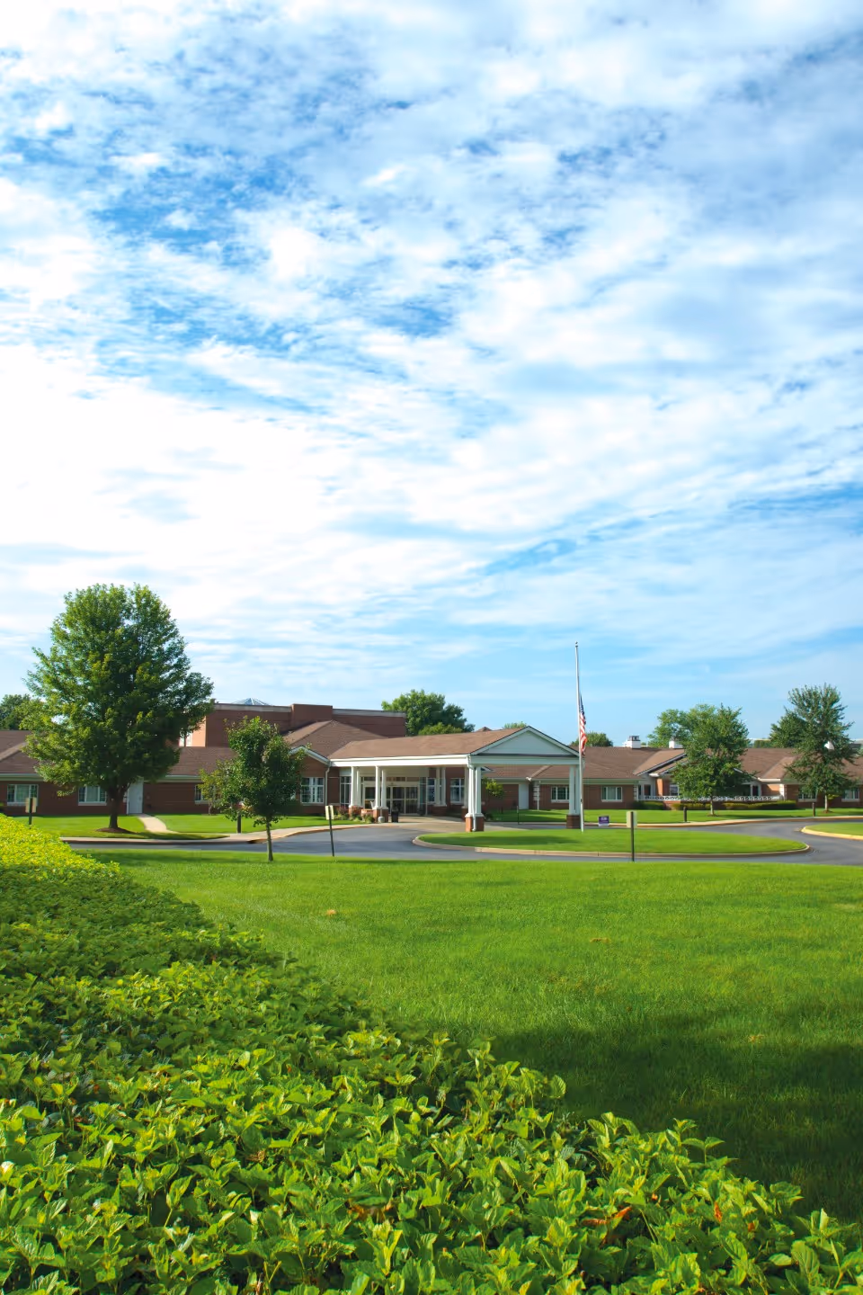 Exterior view of Episcopal Church Home, a single-story brick building with a covered entrance, surrounded by green lawns, trees, and bushes under a partly cloudy blue sky.
