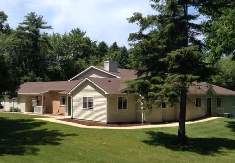 A single-story beige senior living facility with a brown roof, surrounded by green lawn and trees.