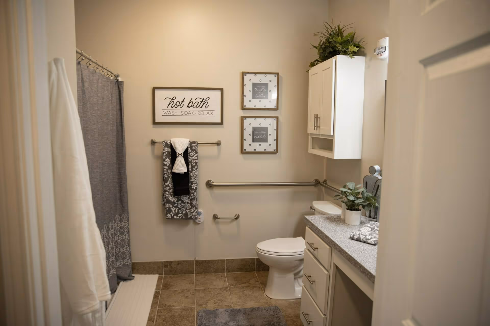 A clean and well-decorated bathroom featuring a toilet, a countertop with a small plant, a white cabinet mounted on the wall, and a shower area with a gray curtain. The walls have three framed signs, one reading 'hot bath WASH SOAK RELAX'. There are grab bars installed near the toilet and along the wall for accessibility, and towels are neatly hung on a towel rack.