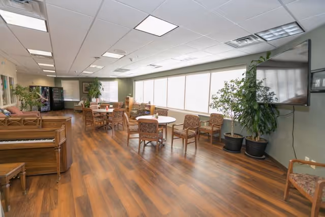 A spacious common area with wooden flooring, several round tables surrounded by cushioned chairs, large windows letting in natural light, a piano on the left side, potted plants, a wall-mounted television, and vending machines in the background.