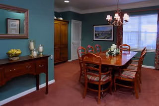 A dining room with a wooden table surrounded by eight wooden chairs with patterned cushions. The walls are painted green and decorated with a framed picture and a mirror. A chandelier with five lampshades hangs above the table, which has a floral centerpiece. There is a wooden side table with decorative items including a bowl of lemons and two candle holders. A window with blinds and patterned curtains lets in natural light.