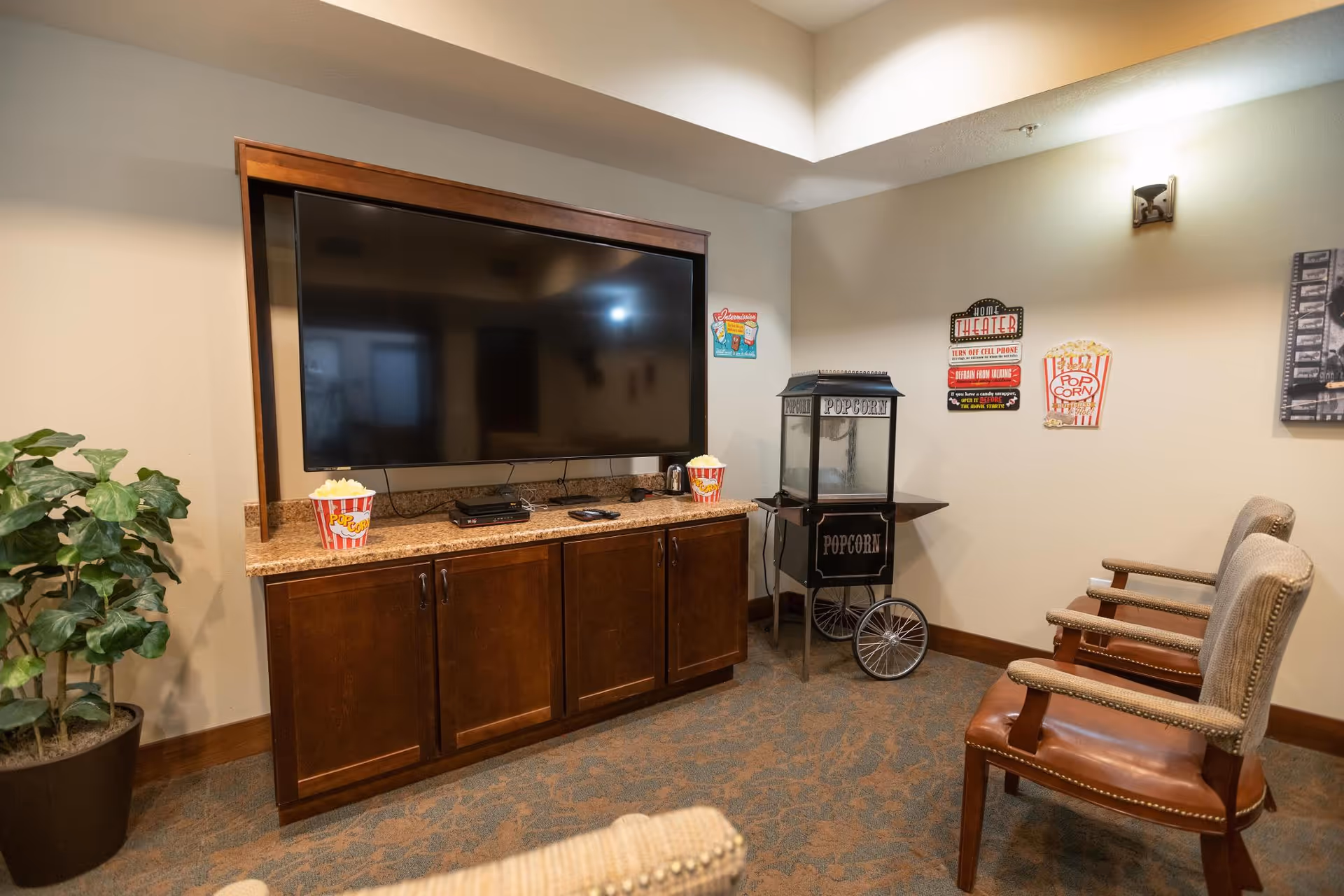 A cozy entertainment room with a large flat-screen TV mounted on a wooden cabinet. There are two popcorn containers on the cabinet and a vintage-style popcorn machine on a small table next to it. Two upholstered chairs with wooden armrests face the TV. The walls have movie-themed decorations, and there is a potted plant in the corner.