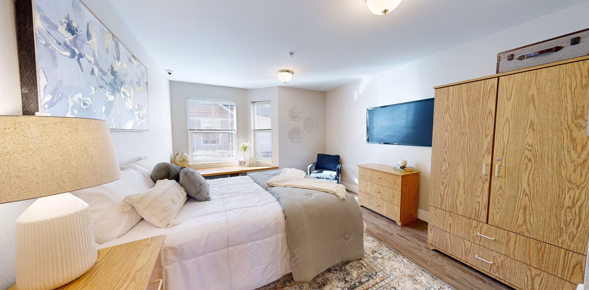 A bright bedroom with a large bed covered in white and gray bedding, a wooden nightstand with a lamp, a wooden dresser and wardrobe, a wall-mounted flat-screen TV, a blue armchair, and two windows letting in natural light. The walls are light-colored and decorated with a painting and circular wall art.