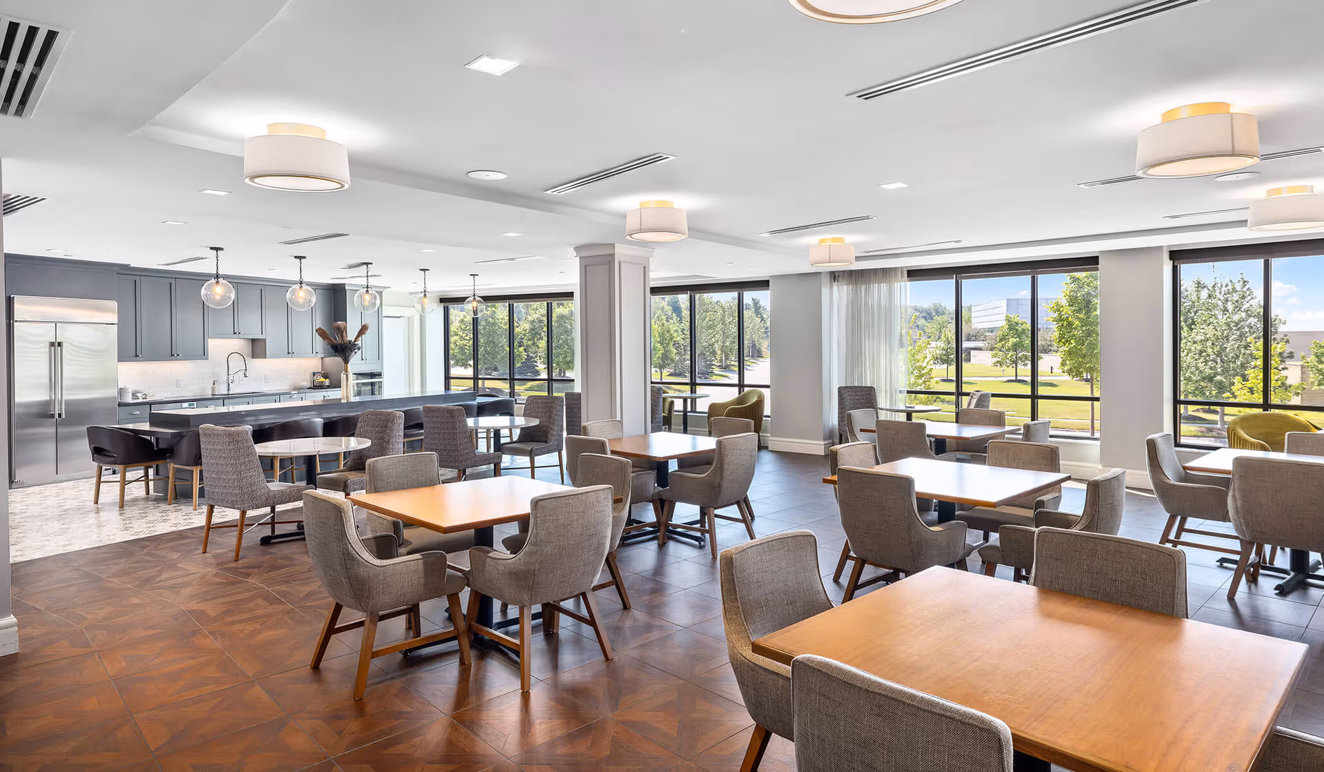 A bright and spacious dining area with multiple wooden tables and gray upholstered chairs. Large windows allow natural light to fill the room and offer views of green trees outside. In the background, there is a modern kitchen with gray cabinets, a stainless steel refrigerator, and a long island with bar stools. The ceiling features several round light fixtures.