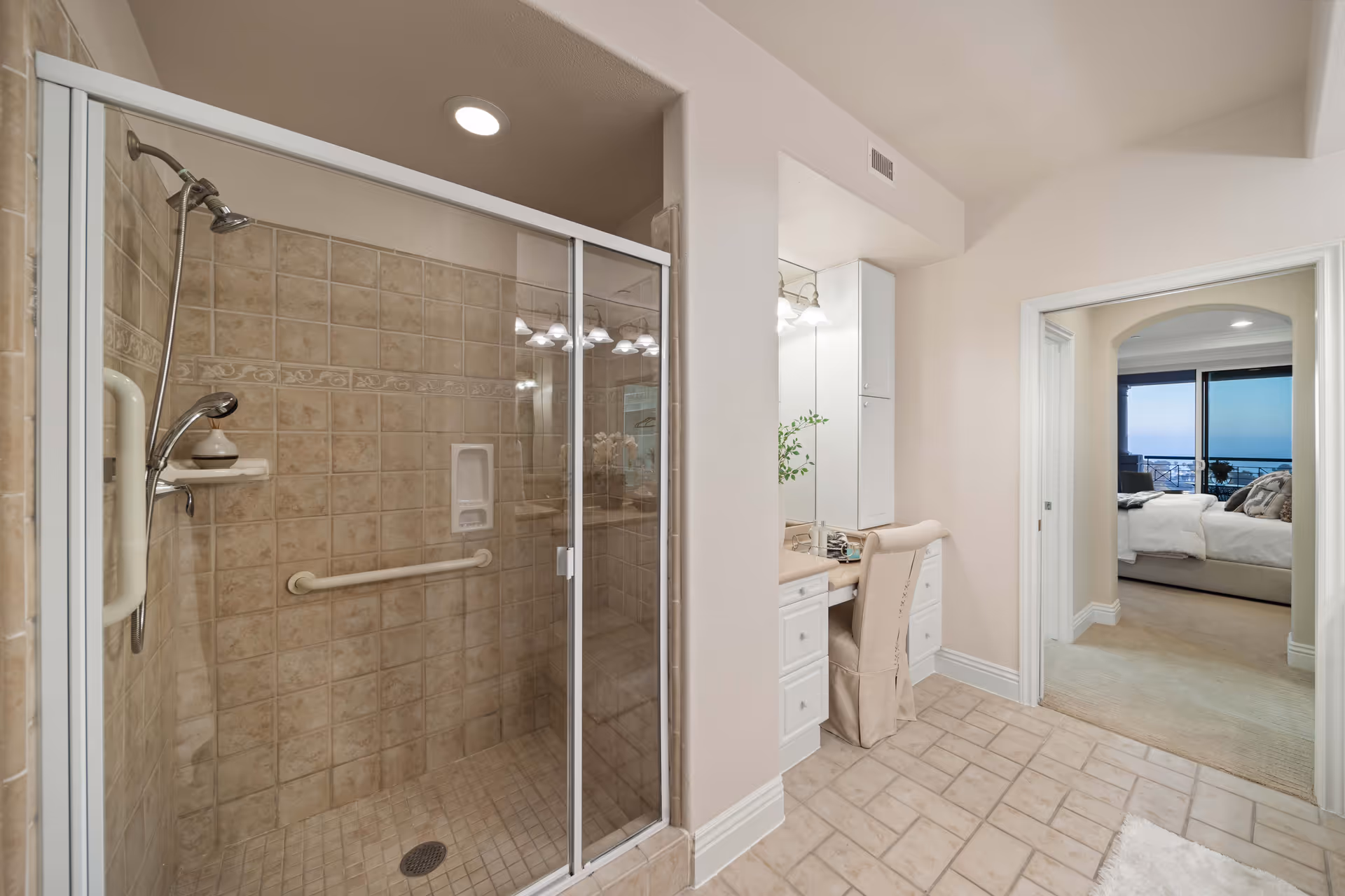 Interior view of a bathroom with a glass-enclosed tiled shower on the left, a vanity area with a chair and mirror in the center, and a doorway leading to a bedroom with a bed and large windows showing an ocean view.