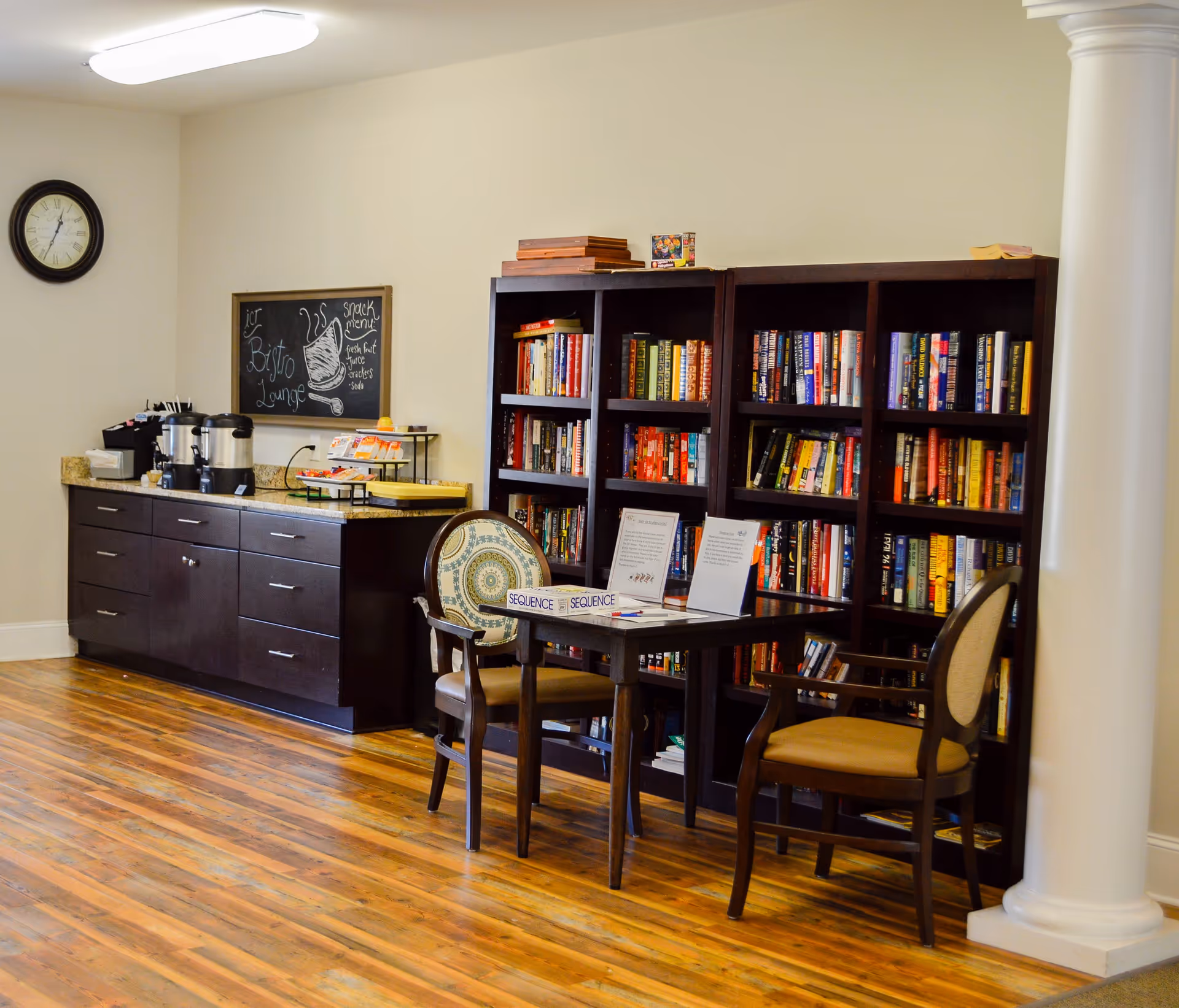 A cozy lounge area with wooden bookshelves filled with books, a small table with two chairs, and a cabinet with coffee dispensers and snacks. A chalkboard on the wall reads 'Bistro Lounge' and lists a snack menu including fresh fruit, juice, crackers, and soda. A clock is mounted on the wall above the cabinet.