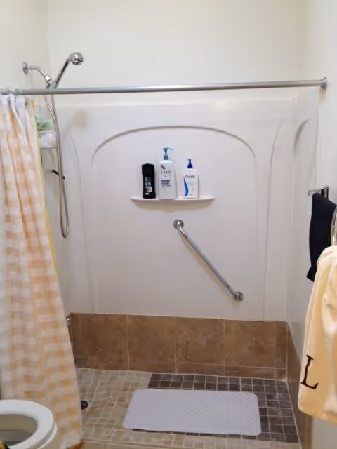 A bathroom shower area with a beige and white checkered shower curtain partially open. Inside the shower, there is a built-in shelf holding three bottles of shower products. A metal grab bar is installed diagonally on the shower wall. The shower floor has small square tiles in shades of brown and beige, with a white bath mat placed on it. A towel rack with a dark towel and a beige towel with the letter 'L' embroidered on it is visible on the right side.