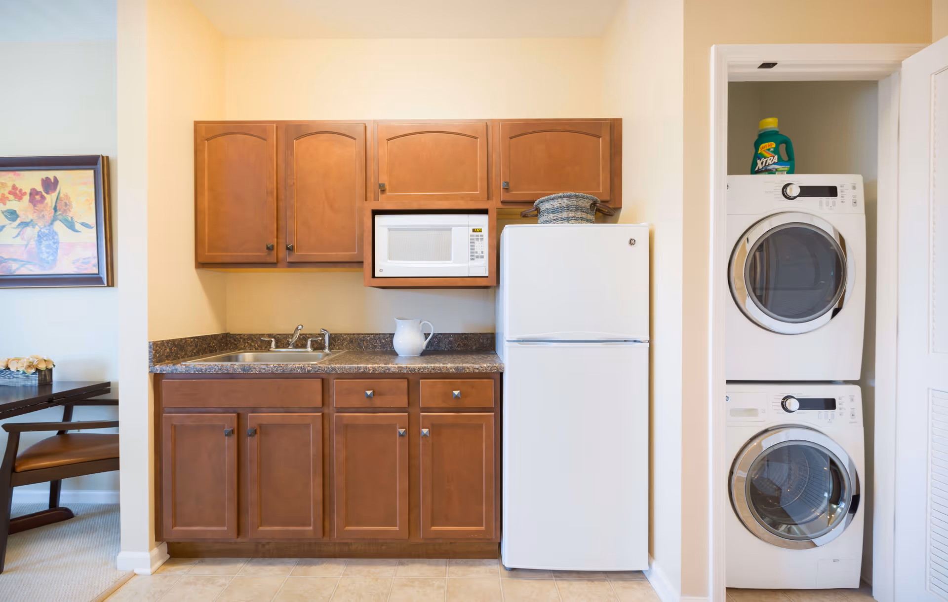 A compact kitchen area with wooden cabinets, a white microwave, a white refrigerator, and a countertop with a sink and a white pitcher. To the right, there is a stacked washer and dryer with a bottle of laundry detergent on top. On the left side, part of a dining table and a framed floral painting are visible.