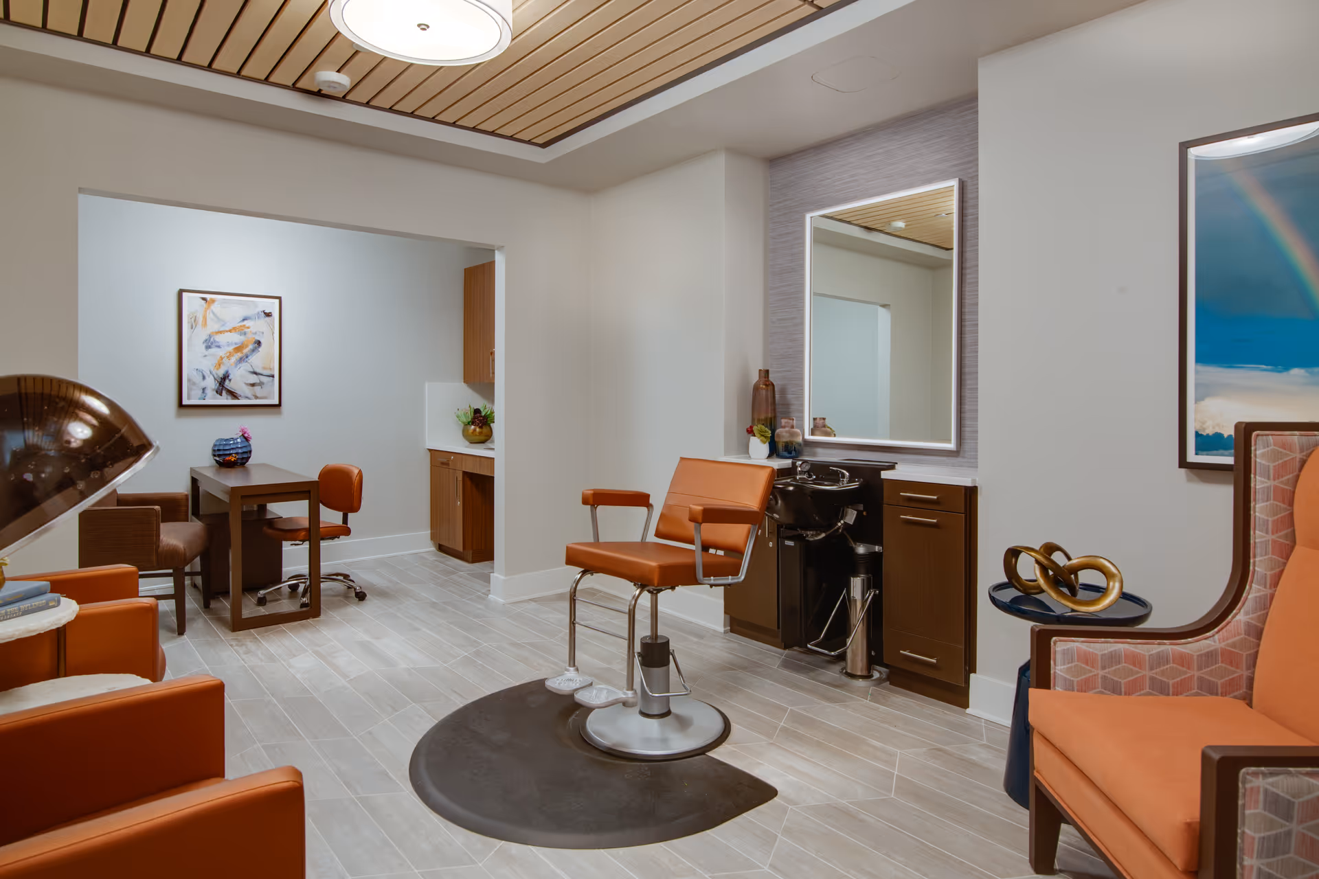 A modern salon area in a senior living facility with an orange salon chair in front of a mirror and sink, surrounded by comfortable seating including an orange armchair and a patterned armchair. The room features light-colored tiled flooring, a wooden ceiling panel, and decorative artwork on the walls. A small table with a decorative item is also visible.