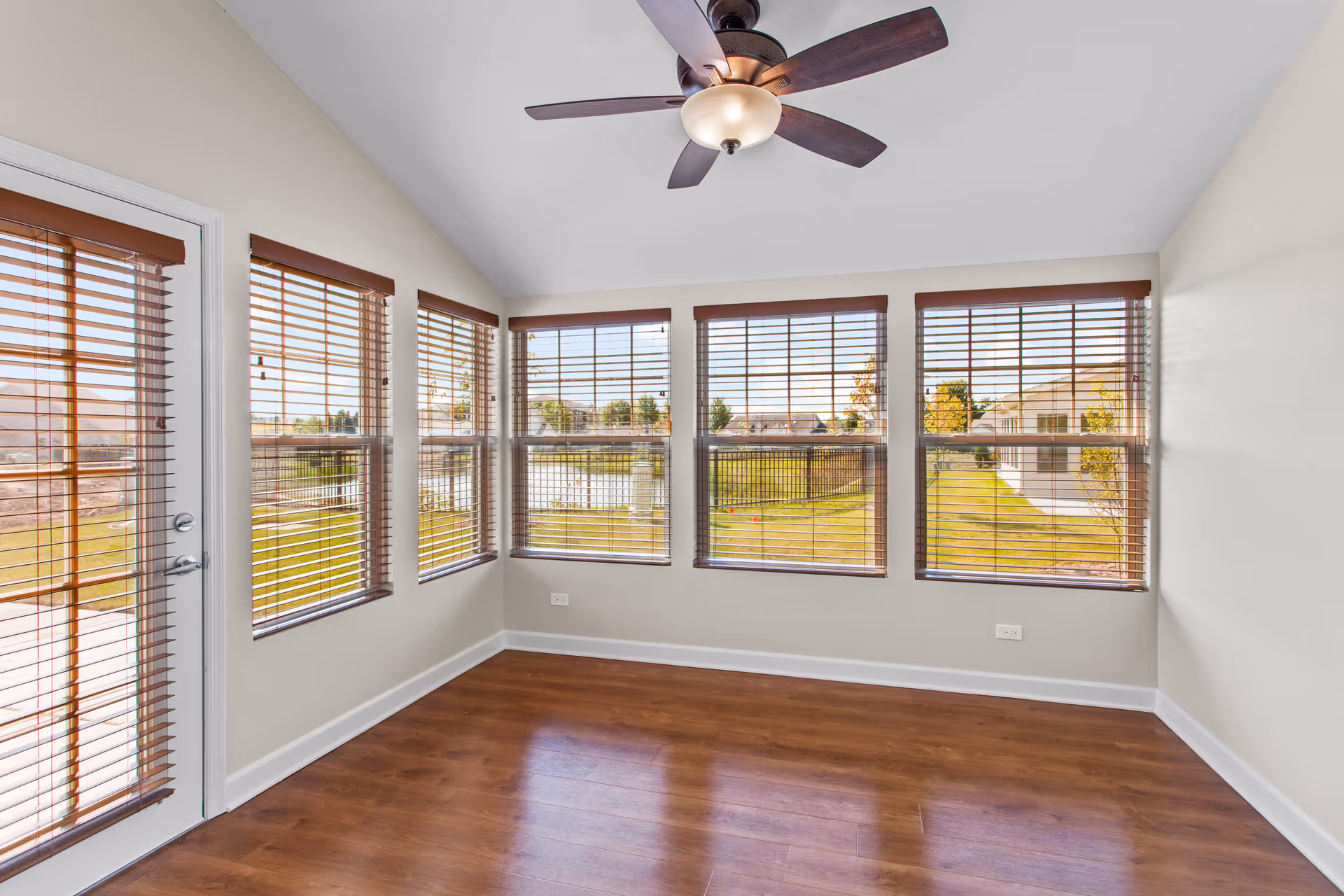 Empty room with wooden floor and multiple large windows with wooden blinds, a ceiling fan with light fixture, and a glass door leading outside. The room is bright with natural light and has beige walls.