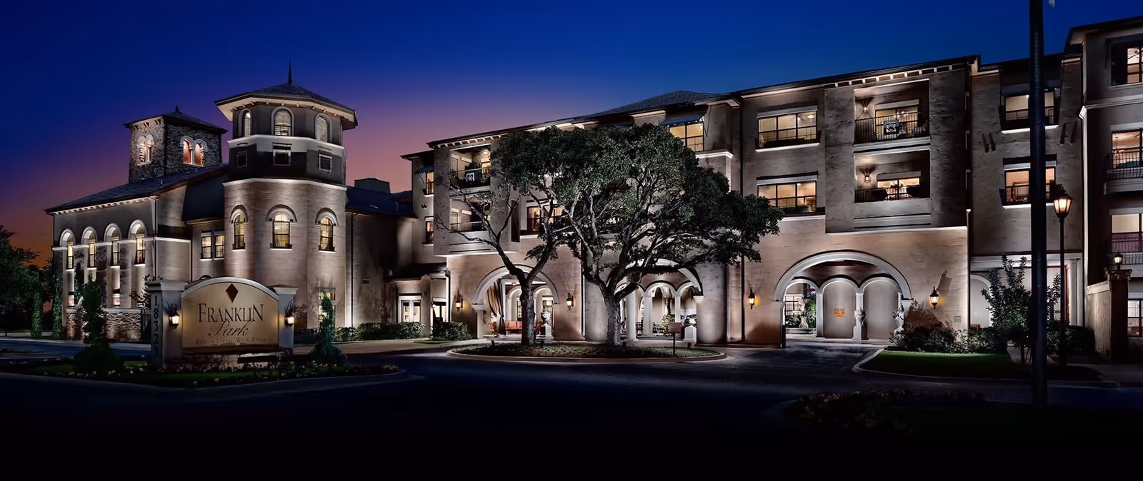 Exterior nighttime view of Franklin Park Sonterra, a large, well-lit senior living facility with a multi-story building featuring arched windows and entrances, a prominent tree in front, and a sign displaying the facility's name.