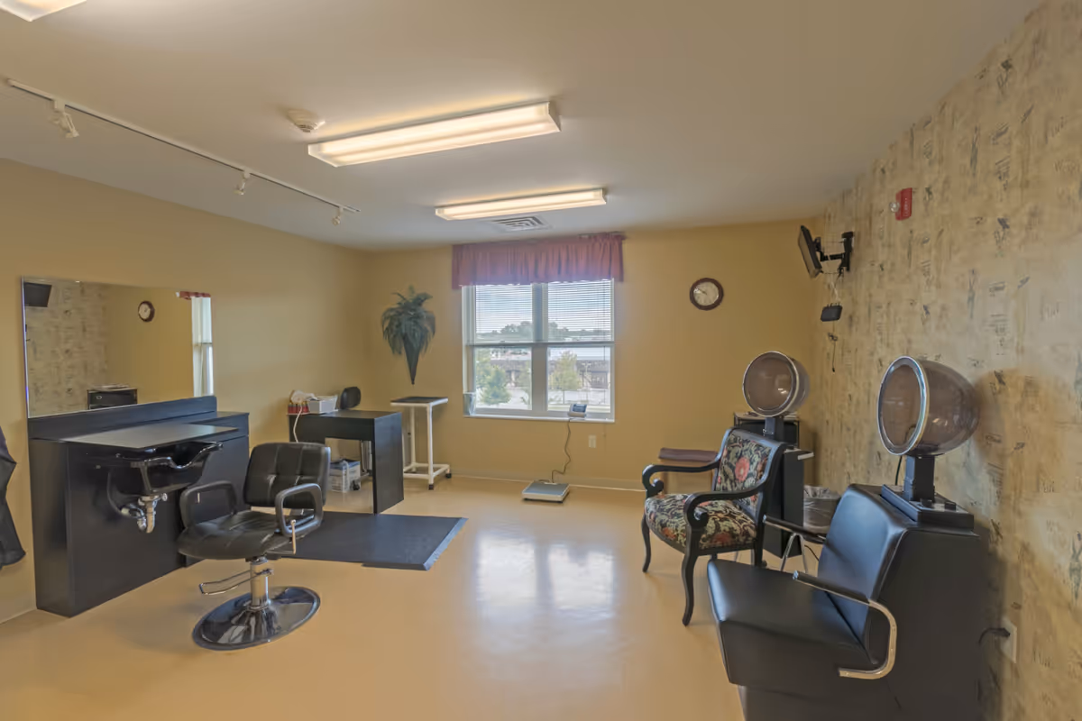 Interior view of a hair salon room in a senior living facility with a black salon chair in front of a sink and mirror, two vintage hair dryers with chairs underneath, a floral upholstered chair, a window with blinds and a red valance, a wall clock, and a small desk with a plant on the wall.