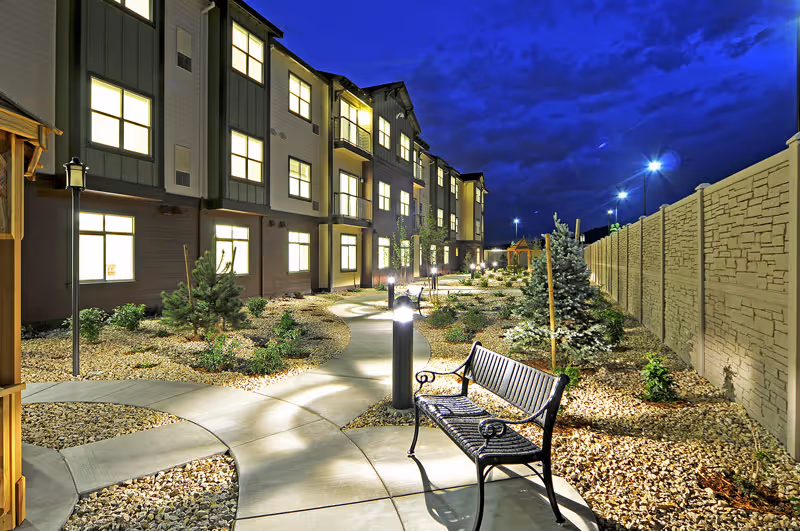 Night view of an outdoor garden area at a senior living facility with a winding concrete pathway, illuminated lamp posts, benches, small trees, and shrubs. The building with multiple lit windows is visible on the left side, and a tall stone-patterned wall runs along the right side.