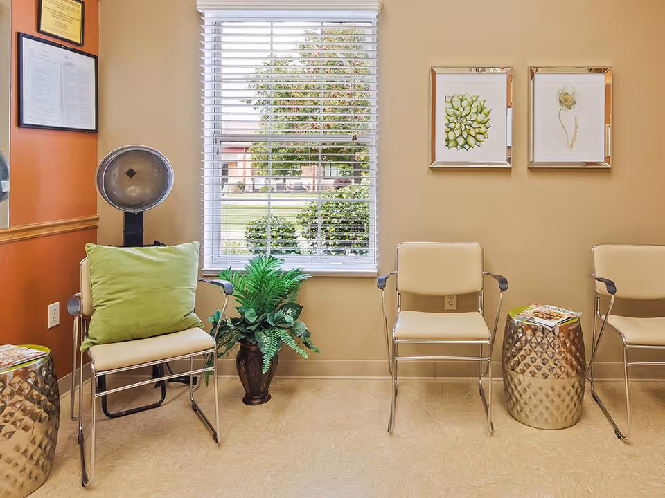 Bright waiting area with three beige chairs, a green pillow and potted plant by a window, framed botanical prints and metallic side tables.