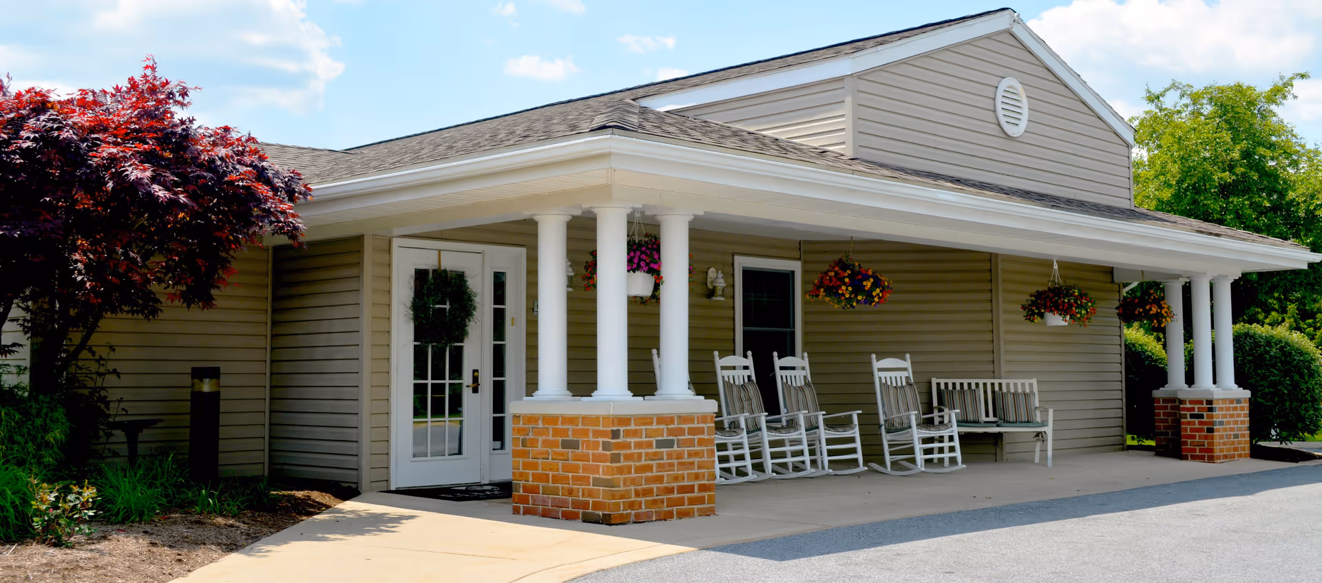 Exterior view of a single-story building with beige siding and a covered porch supported by white columns with brick bases. The porch has several white rocking chairs and hanging flower baskets. There is a glass door with a wreath and a small window next to it. The surrounding area includes a small tree with red leaves and green bushes under a partly cloudy sky.
