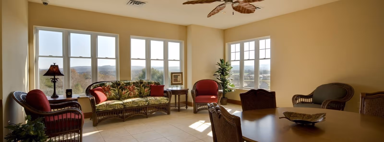 A bright living room with large windows letting in natural light and offering a view of trees and hills outside. The room features wicker furniture including a sofa with floral cushions, two armchairs with red cushions, a side table with a lamp, a potted plant, and a wooden dining table with chairs. The walls are painted a warm beige color and the floor is tiled.