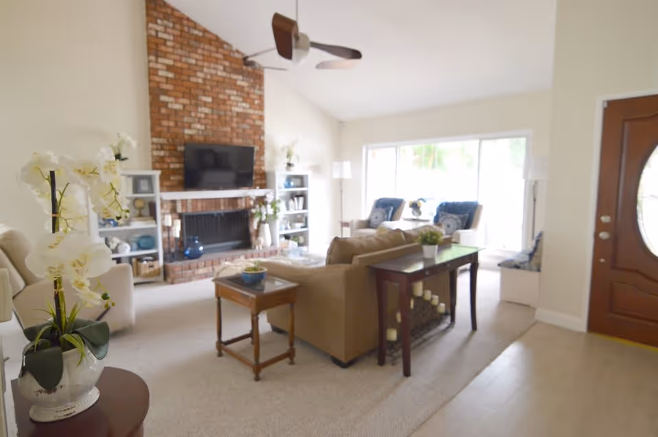 A bright living room with a beige sofa, wooden side tables, and a brick fireplace with a mounted flat-screen TV. There are two white shelves flanking the fireplace with decorative items, a ceiling fan, and large sliding glass doors letting in natural light. A wooden front door with an oval glass window is visible on the right side.