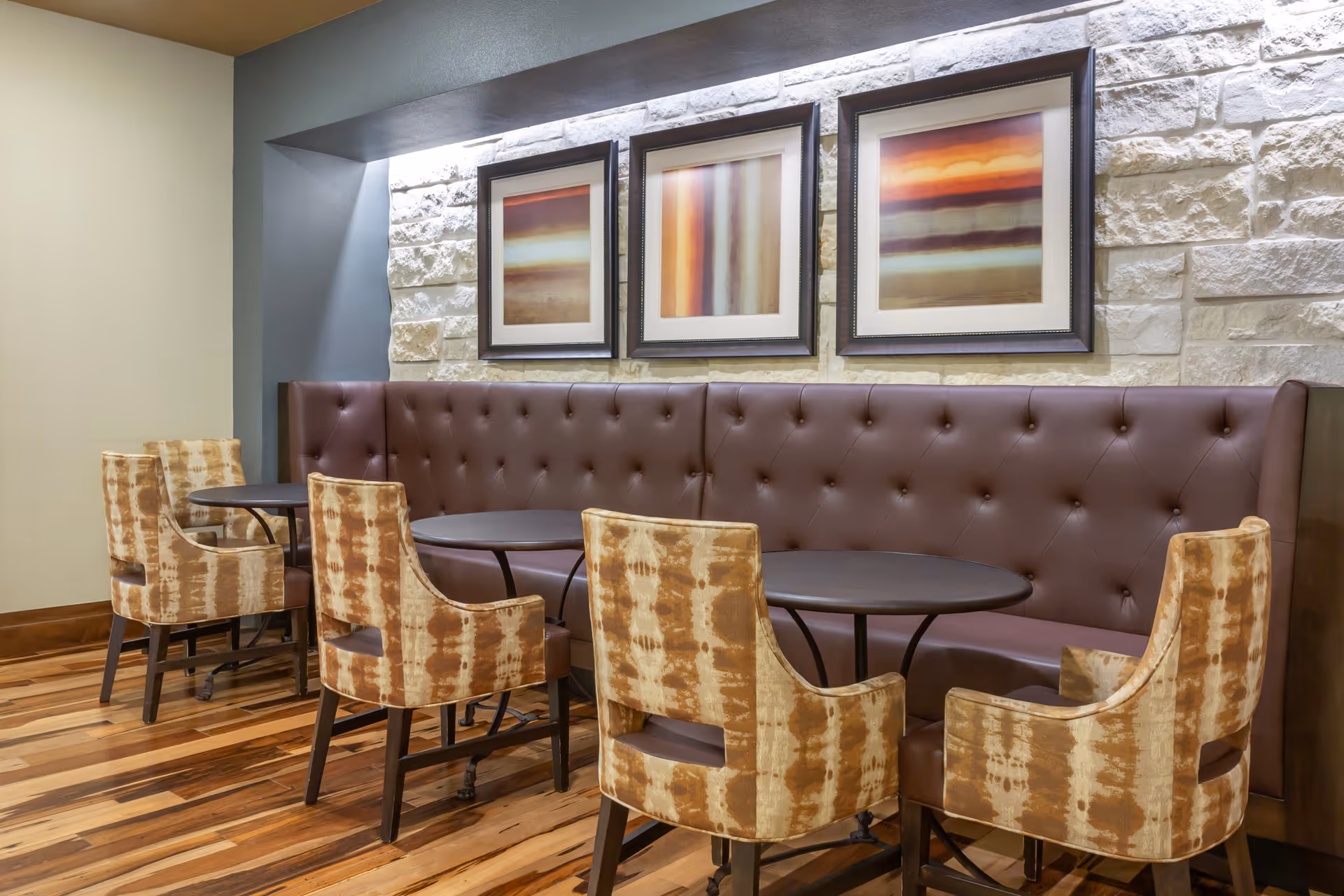 A cozy dining area with a long brown tufted leather bench against a stone wall, three abstract framed paintings above it, and four patterned upholstered chairs around two dark round tables on a wooden floor.