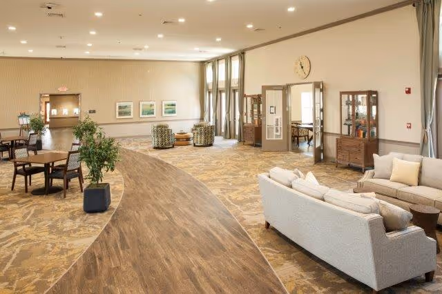A spacious, well-lit common area in a senior living facility featuring a curved wooden floor pathway, beige patterned carpet, multiple seating arrangements including sofas, armchairs, and tables with chairs. Large windows with curtains allow natural light, and there are framed pictures on the walls and a clock above a set of double doors.