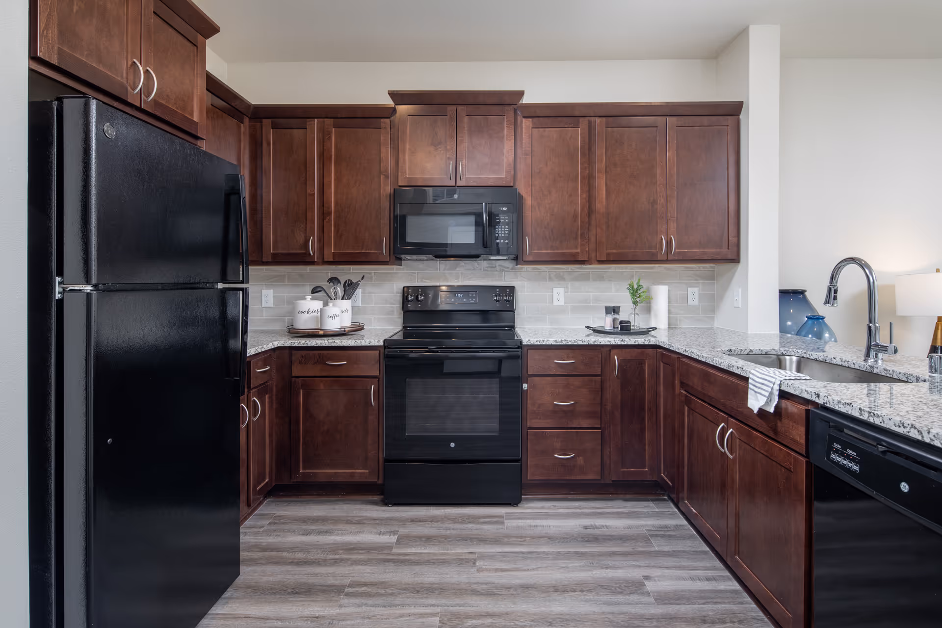 Modern kitchen with dark wood cabinets, black refrigerator, stove, microwave, and dishwasher. Granite countertops with a sink and faucet, light gray tile backsplash, and wood-look flooring.