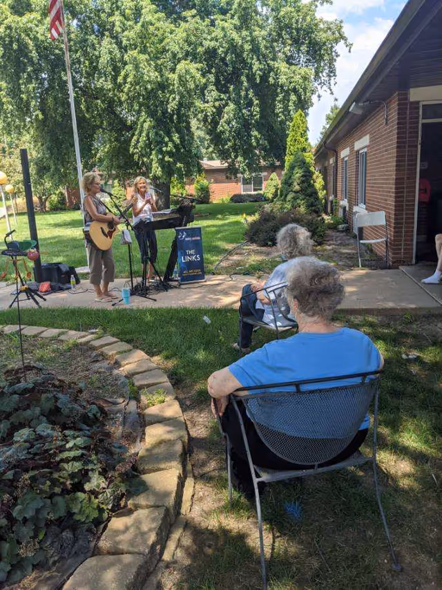 Two women performing music outdoors, one playing a guitar and the other singing into a microphone, with two elderly women seated on metal chairs watching them. The setting is a garden area with green grass, trees, and a brick building in the background under a partly cloudy sky.