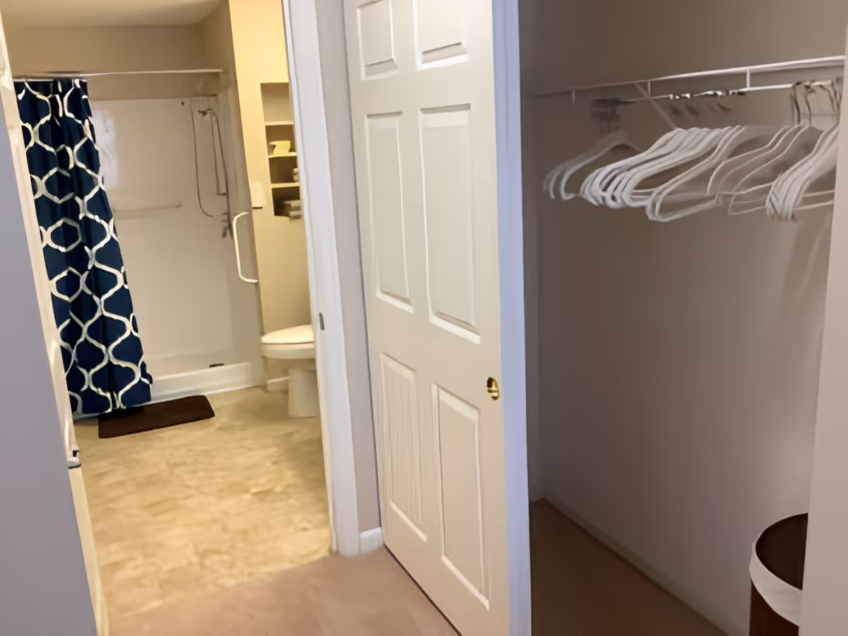 View of a bathroom with a shower area featuring a blue and white patterned shower curtain, a toilet, and shelving with towels. Adjacent to the bathroom is an open closet with white hangers on a rod and a laundry basket on the floor.