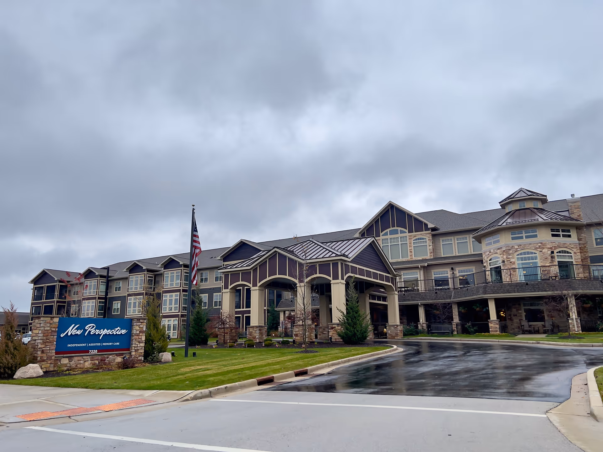 Front entrance and facade of the New Perspective Senior Living building with a driveway, sign, and American flag under a cloudy sky.