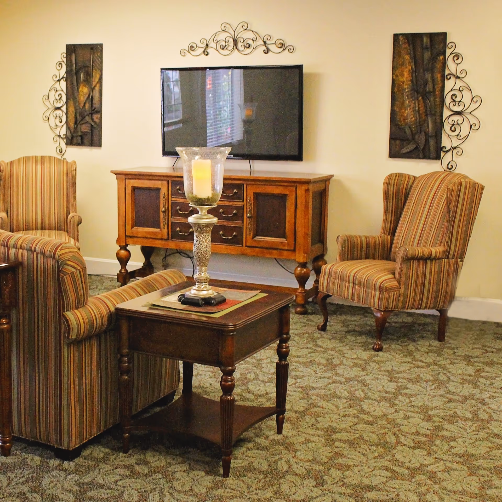 A cozy living room with two striped armchairs and a matching sofa arranged around a wooden coffee table with a decorative candle holder. A wooden TV stand with drawers supports a flat-screen television mounted on the wall, flanked by two decorative metal wall art pieces.