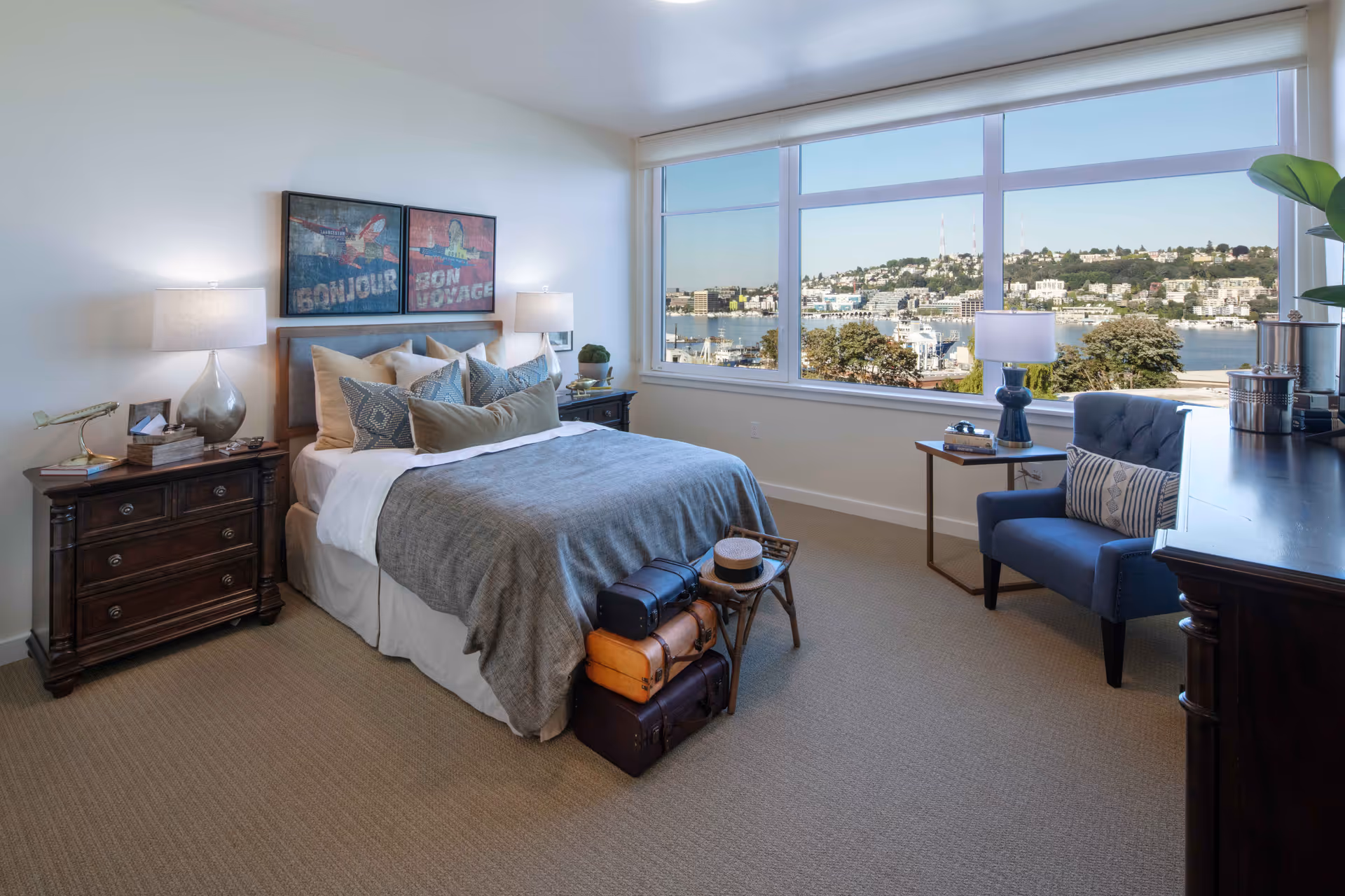 A bright bedroom with a large window offering a scenic view of a waterfront and hillside buildings. The room features a neatly made bed with multiple pillows, two bedside tables with lamps, a blue armchair with a decorative pillow, a small side table with books and a lamp, and a stack of vintage suitcases at the foot of the bed.