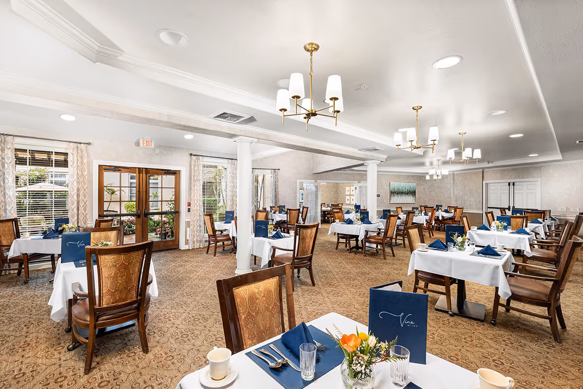 A spacious dining room with multiple tables covered in white tablecloths, each set with blue napkins, glassware, and small floral centerpieces. The room features patterned carpet, elegant wooden chairs with cushioned seats, large windows with curtains, and brass chandeliers hanging from the ceiling. Double glass doors lead outside, and the atmosphere is bright and inviting.