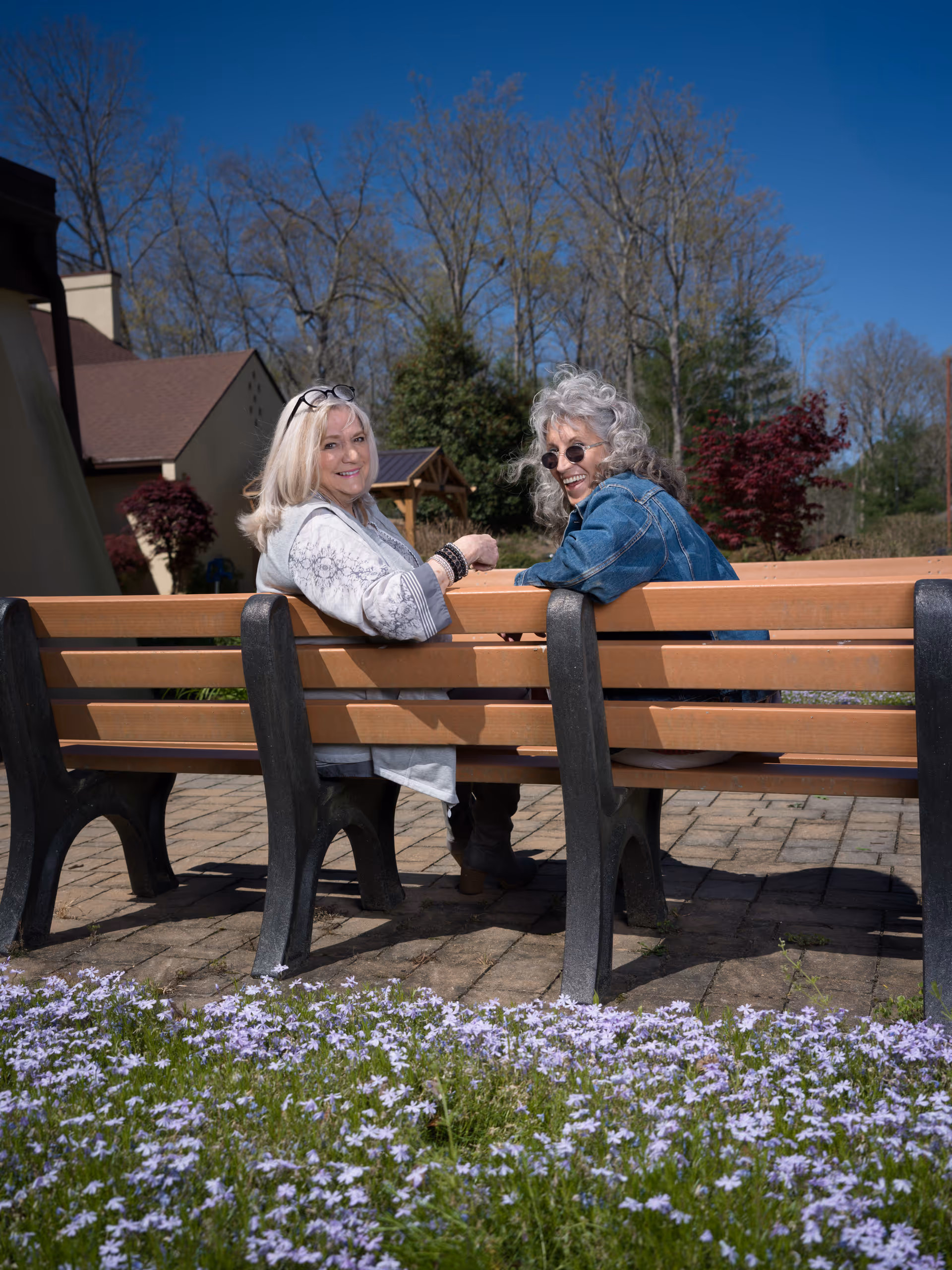 Two older women sitting on a wooden bench outdoors, smiling and looking back towards the camera. They are surrounded by a garden with purple flowers in the foreground and trees in the background under a clear blue sky.
