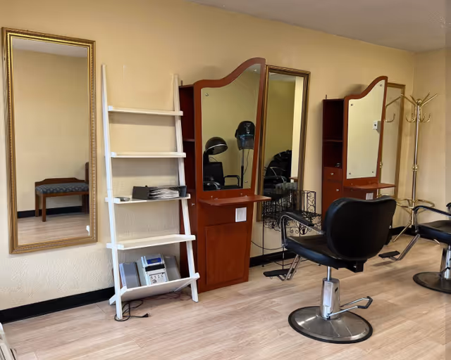 Interior of a salon area with two black salon chairs in front of wooden styling stations with mirrors. There is a white leaning shelf holding papers and a device, a gold-framed wall mirror, and a gold coat rack in the corner. The floor is light wood and the walls are beige.