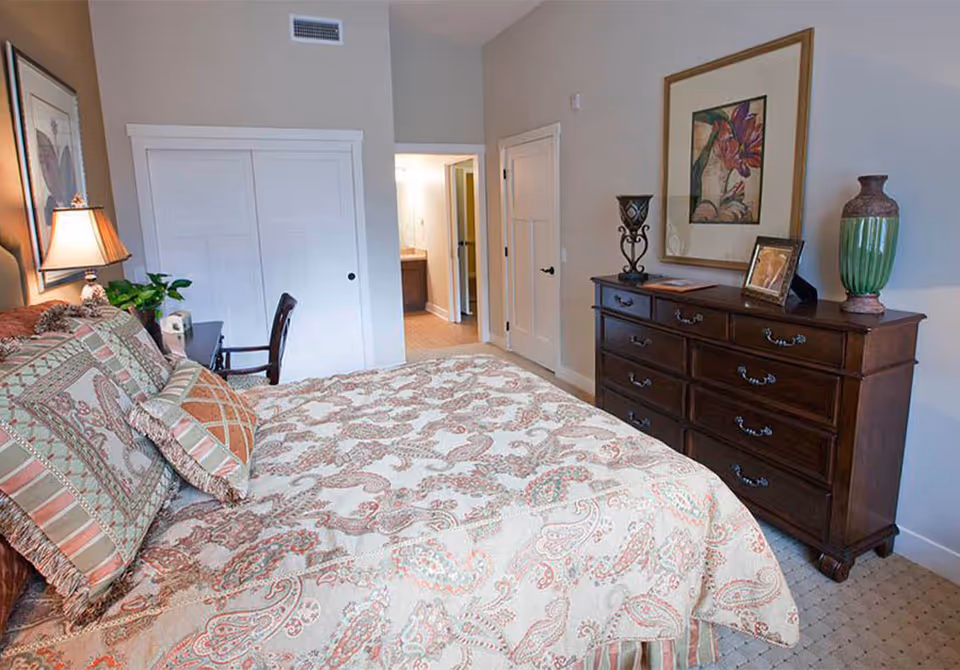 A bedroom with a large bed covered in a patterned quilt and multiple pillows. There is a wooden dresser with decorative items and framed artwork above it. A door leads to a bathroom with a visible sink and mirror. The room has neutral-colored walls and carpeted flooring.