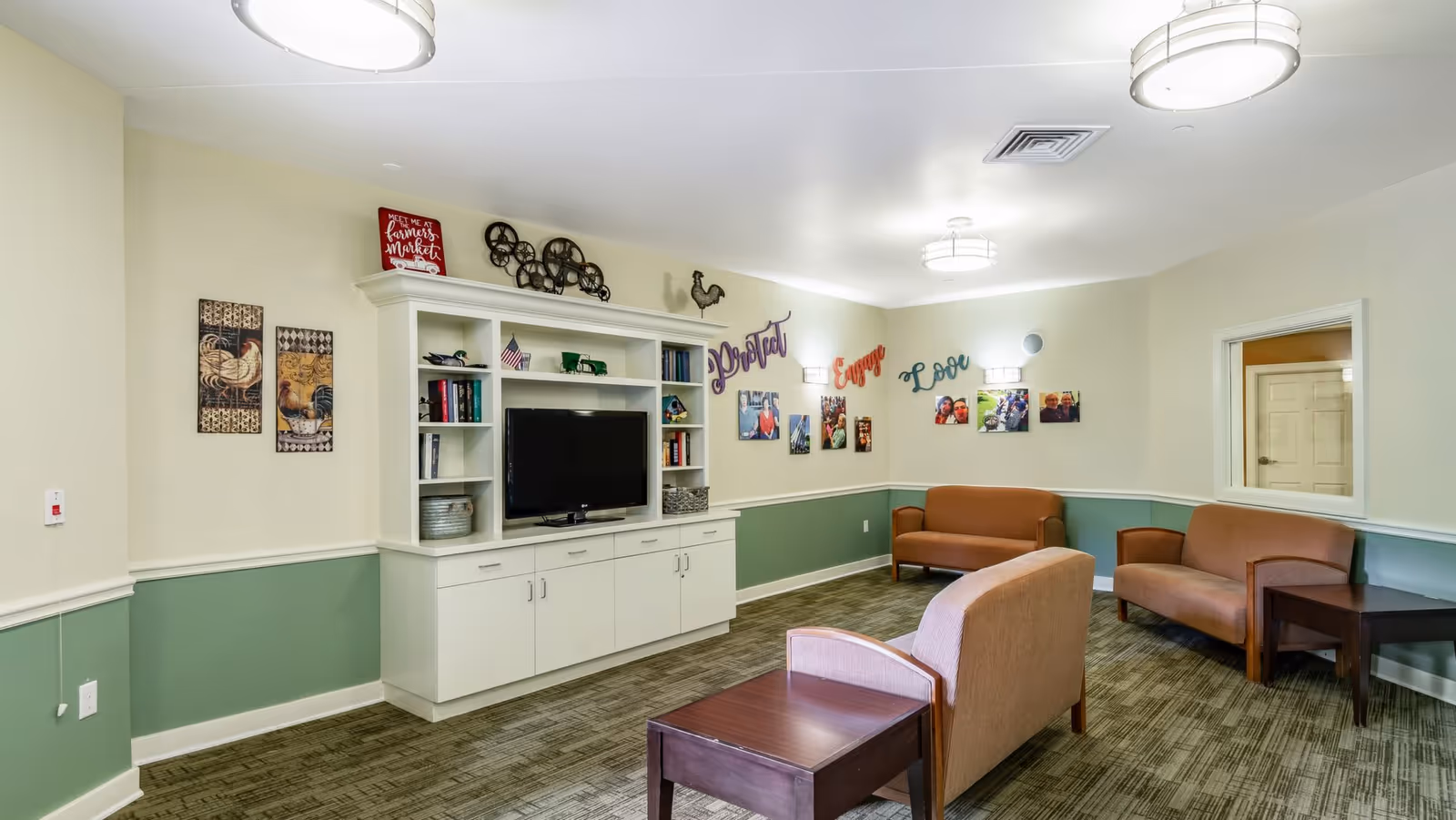 A cozy living room area in Morningside Place with beige and green walls, featuring a white entertainment center with a TV, books, and decorative items. There are three brown upholstered chairs and two brown sofas arranged around the room. The walls are decorated with framed pictures and colorful words 'Protect', 'Engage', and 'Love'. The room is well-lit with ceiling lights.