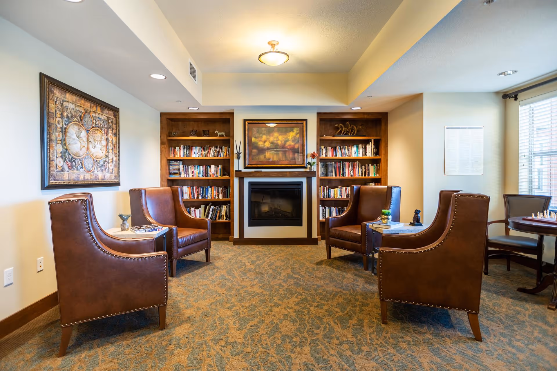 A cozy senior living common area with four brown leather armchairs arranged around two small tables. Behind the chairs is a fireplace flanked by built-in wooden bookshelves filled with books and decorative items. The room has a patterned carpet, a large framed painting above the fireplace, and a window with blinds letting in natural light.