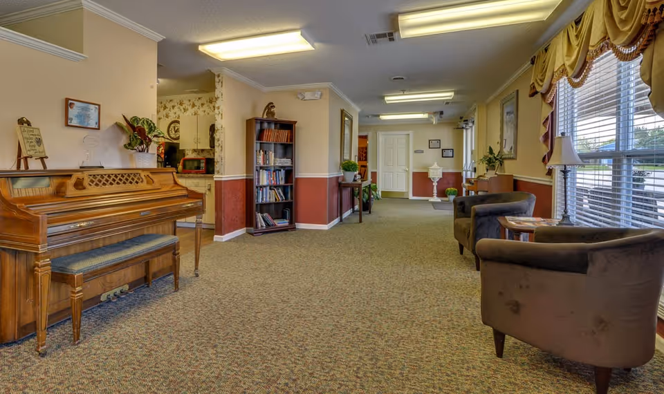 A bright nursing center common room with a wooden piano and bench on the left, bookshelves and plants along the center, and upholstered chairs by large windows on the right.