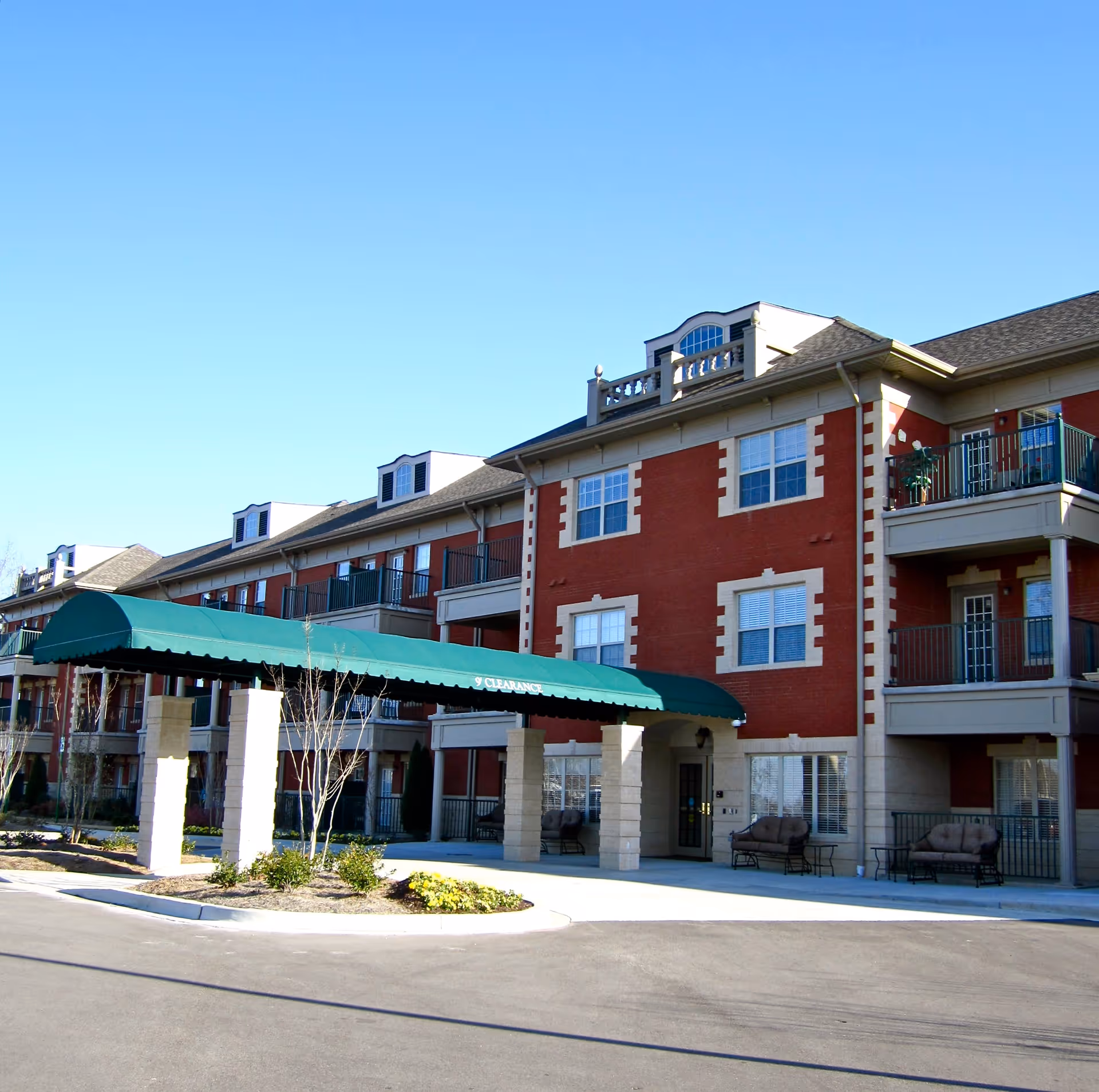 Exterior view of a multi-story senior living community building with red brick and beige accents. The building features balconies, large windows, and a green canopy entrance supported by stone pillars. There are some small landscaped areas with plants and bushes near the entrance.