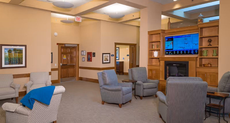 A cozy living room area in a senior living facility with several upholstered armchairs arranged around a wooden entertainment center that houses a flat-screen TV displaying a weather forecast. The room features beige walls, carpeted floor, framed artwork, and wooden doors leading to other rooms. Ceiling lights and an exit sign are visible.