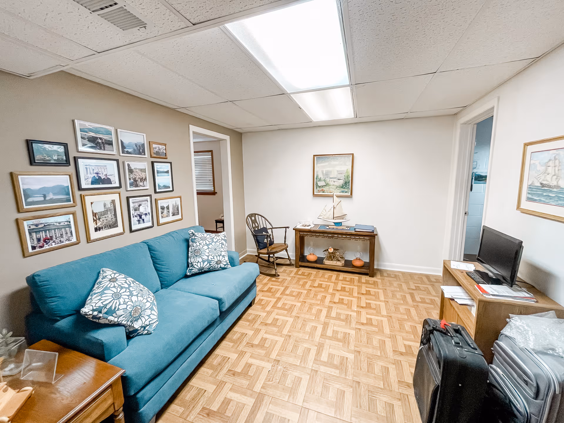 A cozy living room with a teal sofa adorned with floral pillows, a wooden rocking chair, and a wooden console table decorated with a model sailboat and two small pumpkins. The walls are decorated with framed pictures and paintings, and there is a small wooden desk with a computer monitor and some luggage nearby. The floor has a parquet pattern, and the ceiling has fluorescent lighting.