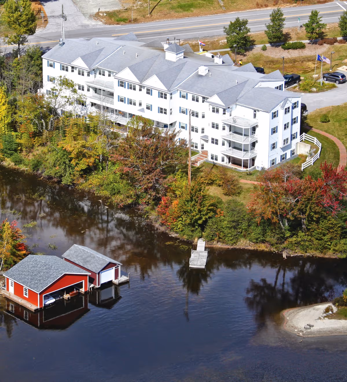 Aerial view of a large white multi-story assisted living facility building next to a calm body of water with two red boathouses on the water. The building is surrounded by trees with autumn foliage and a road runs behind the building.