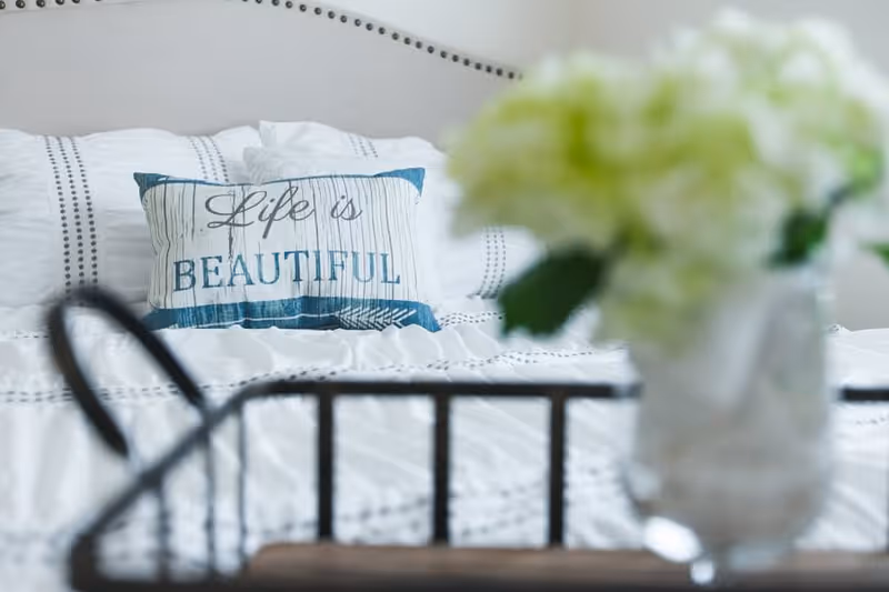 Close-up view of a bed with white bedding and a decorative pillow that reads 'Life is BEAUTIFUL'. In the foreground, there is a blurred vase with white flowers on a black metal tray.
