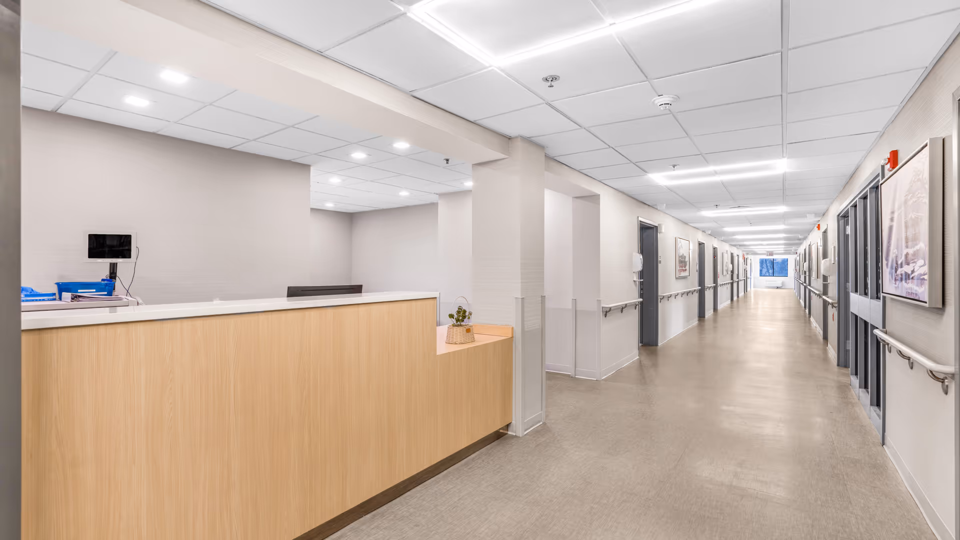 A long, well-lit hallway in a healthcare facility with a light wood reception desk on the left side. The hallway has handrails on both sides, several closed doors, and framed pictures on the walls. The ceiling has recessed lighting and white tiles.