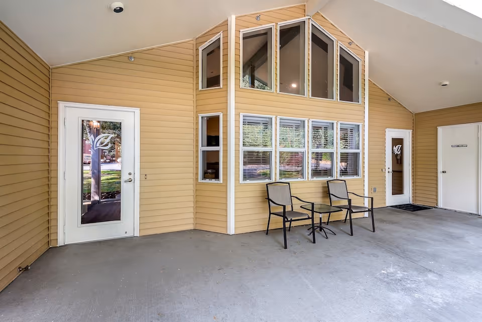Covered outdoor seating area with two chairs and a small glass table in front of a beige building with multiple windows and two white doors, one door has a leaf design on the glass.