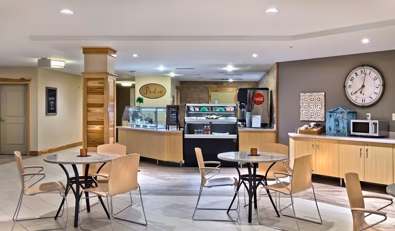 A bright and clean dining area with round tables and wooden chairs. In the background, there is a food service counter labeled 'Bistro' with a display case containing food items and a Coca-Cola beverage dispenser. To the right, there is a countertop with a microwave, a large wall clock, and decorative items on the wall.