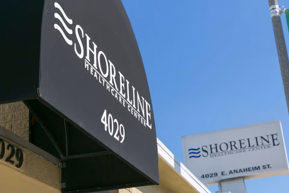 Two signs for Shoreline Healthcare Center, one black awning with white text showing the name and number 4029, and a white freestanding sign with the name, logo, and address 4029 E. Anaheim St. under a clear blue sky.