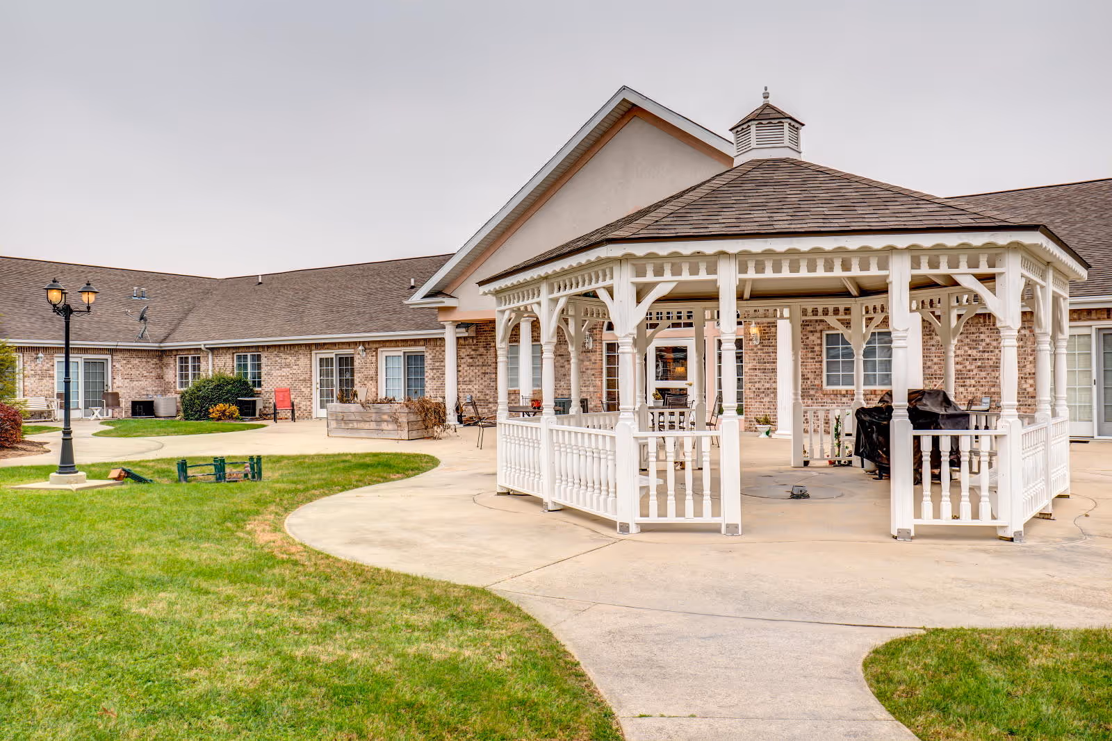 Outdoor area of Brookstone Estates of Charleston featuring a white wooden gazebo on a concrete patio surrounded by green grass, with a brick building in the background and a black street lamp on the left side.