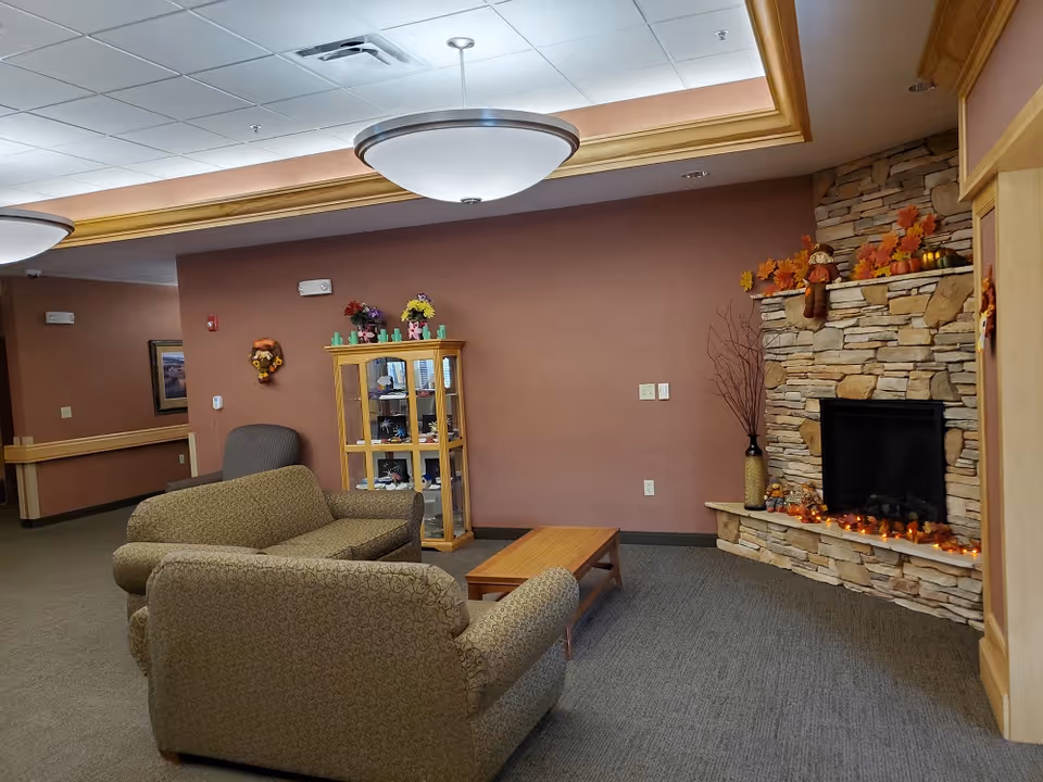 A cozy living room area with two patterned armchairs and a wooden coffee table facing a stone fireplace decorated with autumn leaves and small pumpkins. There is a glass display cabinet with decorative items and a single armchair against a brown wall. The ceiling has recessed lighting and a large round ceiling light fixture.