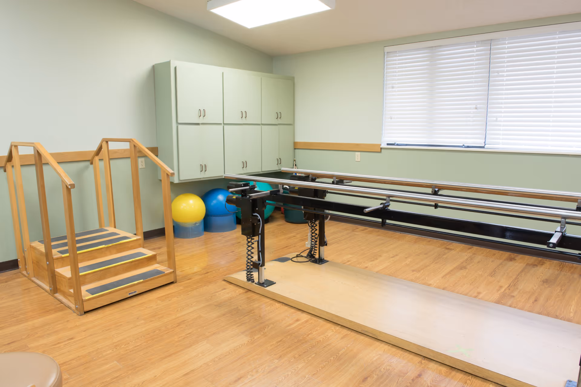 Physical therapy/rehab room with parallel bars, wooden practice stairs, exercise balls, and wall cabinets under a window.